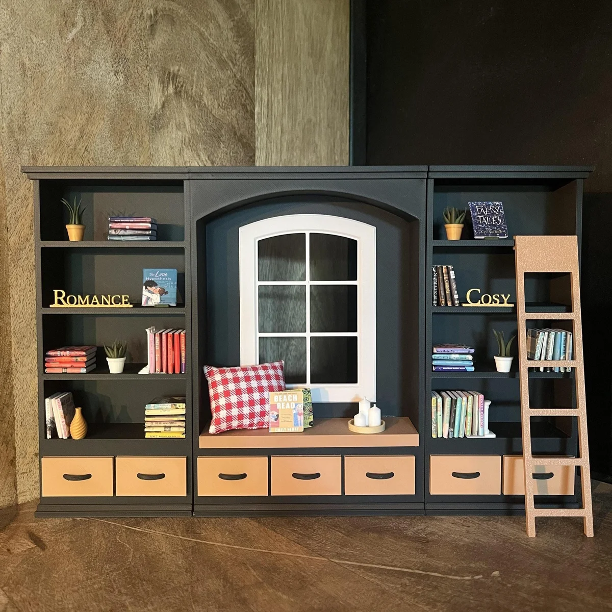 Cozy miniature dollhouse bookcase in black with central window reading nook, cushioned bench, and warm-toned drawers, styled in a rustic 1:12 scale library setting